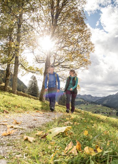 Two hikers walking along a trail in an autumn landscape with mountains, trees, and a cloudy sky, sunlight shining through the trees. | © Kleinwalsertal Tourismus eGen • Photographer: Dominik Berchtold