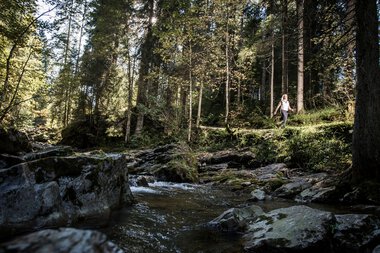 A person hiking along a small stream in a dense forest with tall trees, sunlight filtering through the foliage. | © Kleinwalsertal Tourismus eGen • Photographer: Dominik Berchtold