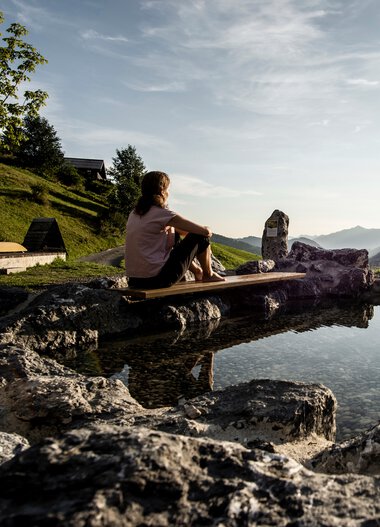 Woman sitting at the Naturerlebnisplatz Rohrweg, surrounded by rocks, overlooking mountains and sunset. | © Kleinwalsertal Tourismus eGen • Photographer: Dominik Berchtold