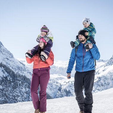 Family winter hiking in the mountains, two children on parents' shoulders, snowy peaks in the background. | © Kleinwalsertal Tourismus eGen • Photographer: Dominik Berchtold