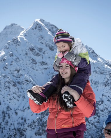 Family winter hiking in the mountains, children on parents' shoulders, with snow-covered peaks in the background. | © Kleinwalsertal Tourismus eGen • Photographer: Dominik Berchtold