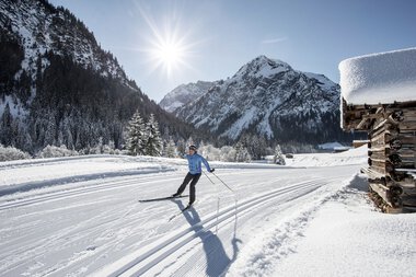 Skifahrer auf Langlaufbahn in verschneiter Winterlandschaft mit Bergen und Sonne im Hintergrund. | © Kleinwalsertal Tourismus eGen • Fotograf: Dominik Berchtold
