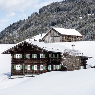 Traditional wooden house in a snowy winter landscape with mountains in the background. | © Kleinwalsertal Tourismus eGen • Photographer: Dominik Berchtold