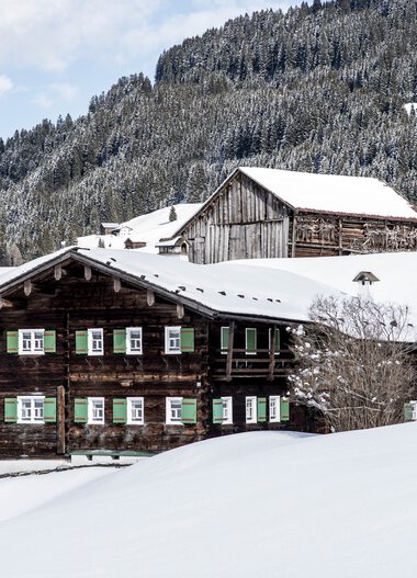 Traditional wooden house in a snowy winter landscape with mountains in the background. | © Kleinwalsertal Tourismus eGen • Photographer: Dominik Berchtold