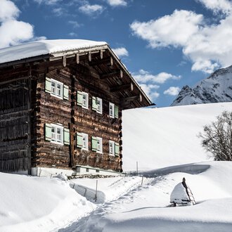 A snowy wooden house in the mountains with a small tree beside it, surrounded by snow, under a cloudy sky. | © Kleinwalsertal Tourismus eGen • Photographer: Dominik Berchtold