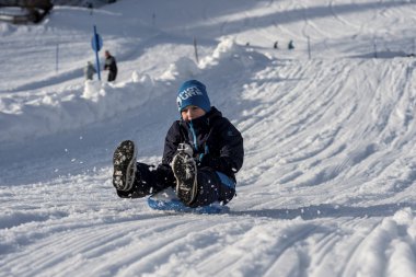 A child is sitting on a sled in the snow, smiling during a winter shoot at Mittelberg, surrounded by snowy mountains. | © Kleinwalsertal Tourismus eGen • Photographer: Dominik Berchtold