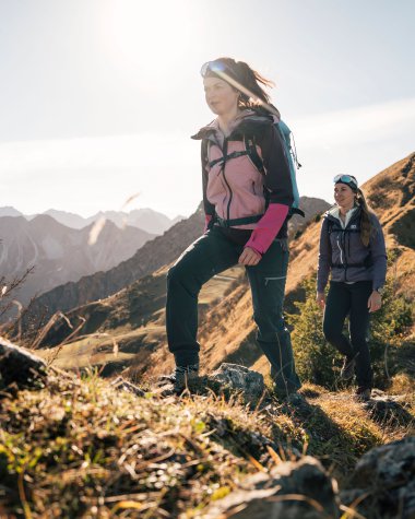 Two women hiking on a mountain trail in the mountains with sunlight, surrounded by nature and rocky terrain.