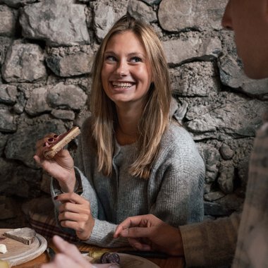 Eine junge Frau mit blonden Haaren lächelt in einer gemütlichen, steinernen Umgebung, während sie mit einem Mann spricht und eine kleine Holzfigur hält. | © Kleinwalsertal Tourismus eGen • Fotograf: Hannah Bichay