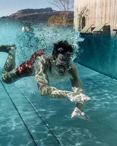 A man is diving into a swimming pool with splashing water, mountains, and a pavilion in the background. | ©  Kleinwalsertal Tourismus eGen • Photographer: Hannah Bichay