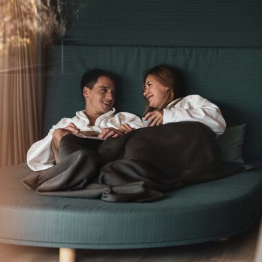 Ein Mann und eine Frau sitzen bequem auf einer orangefarbenen Couch, umhüllt von einer Decke, und lachen miteinander in einem gemütlichen Raum. | © Kleinwalsertal Tourismus eGen • Fotograf: Hannah Bichay