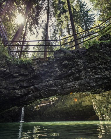 Natural rock arch over water inside a cave, sunlight shines through treetops, wooden bridge in the background. | © Kleinwalsertal Tourismus eGen • Photographer: Oliver Farys