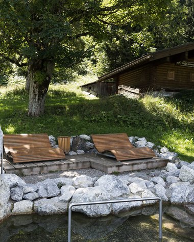 Natural experience area with a foot bath and rocks, surrounded by trees and a small wooden house in the background. | © Kleinwalsertal Tourismus eGen • Photographer: Oliver Farys