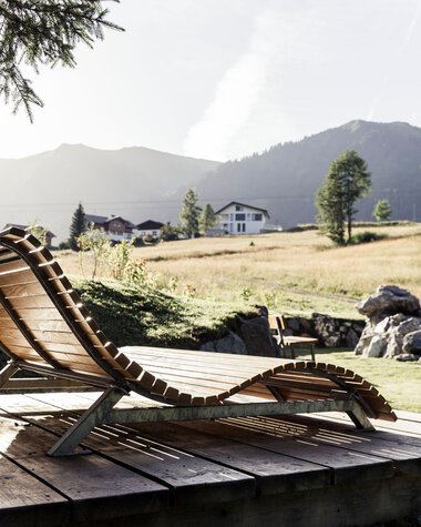 Relaxing wooden lounge chair on a wooden deck in a natural mountain landscape with meadows, trees, and mountains in the background. | © Kleinwalsertal Tourismus eGen • Photographer: Oliver Farys