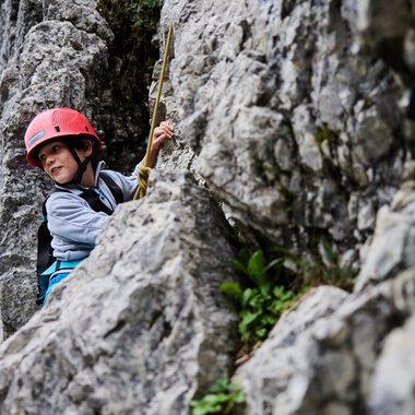 Junge beim Klettertraining an einer Felswand, trägt Helm und Klettergurt, mit Blick nach oben, in einer natürlichen Umgebung. | © Kleinwalsertal Tourismus eGen • Fotograf: Oliver Farys
