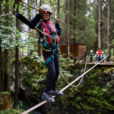 Woman crossing a rope bridge in a forest adventure park, wearing a helmet and safety gear, surrounded by trees. | © Kleinwalsertal Tourismus eGen • Photographer: Oliver Farys