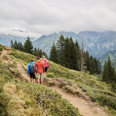 Gruppen von Wanderern unterwegs auf einem Weg in den Bergen, umgeben von Tannenbäumen und aufziehendem Himmel, im Kleinwalsertal. | © Kleinwalsertal Tourismus eGen • Fotograf: Oliver Farys
