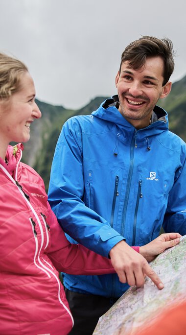 Two hikers with backpacks, laughing and chatting during an autumn hike in Kleinwalsertal, with mountain scenery in the background. | © Kleinwalsertal Tourismus eGen • Photographer: Oliver Farys