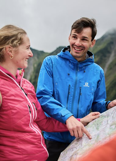 Two hikers with backpacks, laughing and chatting during an autumn hike in Kleinwalsertal, with mountain scenery in the background. | © Kleinwalsertal Tourismus eGen • Photographer: Oliver Farys