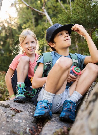 Zwei Kinder auf einem Felsen in einem Wald, eines schaut nach oben, das andere lächelt, Umgeben von Bäumen und Natur. | © Kleinwalsertal Tourismus eGen • Fotograf: Oliver Farys
