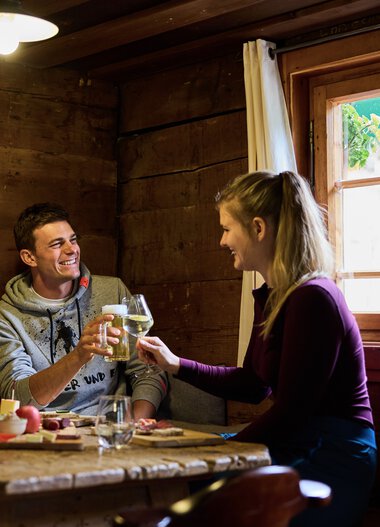 Two people sitting inside a wooden cabin, laughing and clinking wine glasses during a meal. | © Kleinwalsertal Tourismus eGen • Photographer: Oliver Farys
