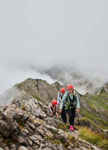 Menschen beim Klettersteig in den Bergen, umgeben von Wolken und Felsen, mit Bergschule Walter Walser-Logo im Bild. | © Kleinwalsertal Tourismus eGen • Fotograf: Oliver Farys