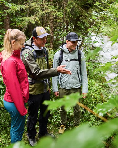 Drei Personen, darunter ein Ranger, führen eine outdoor-Infosession im grünen Wald am Fluss durch. | ©  Kleinwalsertal Tourismus eGen • Fotograf: Oliver Farys