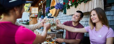 People enjoying summer outdoor dining on a terrasse with wooden houses and colorful flowers, sharing toasts and snacks. | © Kleinwalsertal Tourismus eGen • Photographer: Oliver Farys