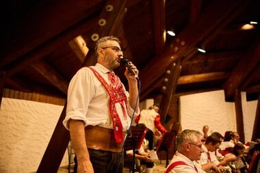 Man in traditional attire singing and performing music at a summer event of the Trachtenkapelle Mittelberg in a wooden hall. | © Kleinwalsertal Tourismus eGen • Photographer: Oliver Farys