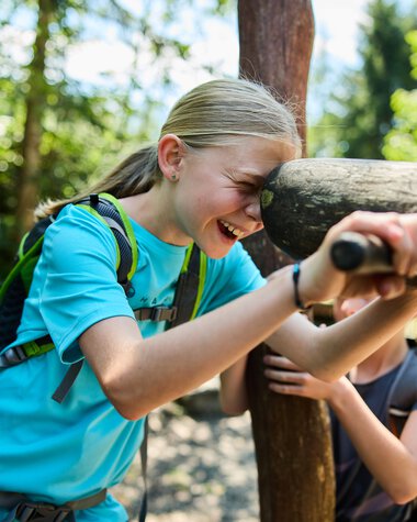 Smiling young girl playing in the forest, hitting a log with her head. | © Kleinwalsertal Tourismus eGen • Photographer: Oliver Farys