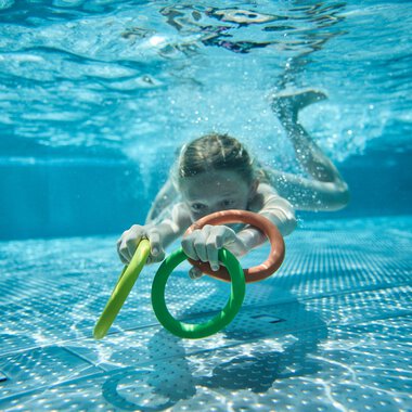 Ein junges Mädchen schwimmt im Freibad, hält sich an bunten Wasserhölzern und schaut nach vorne, während sie unter Wasser ist. | © Kleinwalsertal Tourismus eGen • Fotograf: Oliver Farys