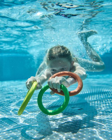A young girl swimming in an outdoor pool, holding colorful foam noodles and looking forward underwater. | © Kleinwalsertal Tourismus eGen • Photographer: Oliver Farys