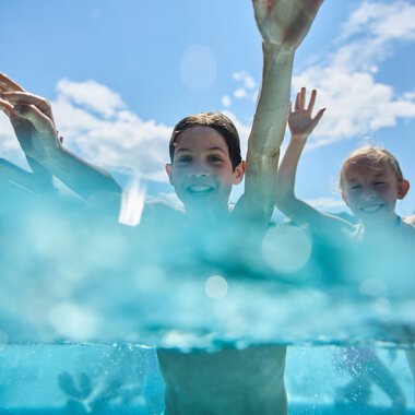 Two children swimming and smiling in the outdoor pool at Freibad Riezlern, with a bright blue sky and clouds in the background. | © Kleinwalsertal Tourismus eGen • Photographer: Oliver Farys