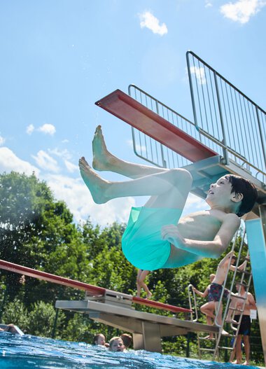 A boy jumps from a diving board into an outdoor swimming pool in summer, with other swimmers and green mountains in the background. | © Kleinwalsertal Tourismus eGen • Photographer: Oliver Farys