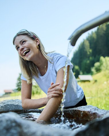 Woman sitting at the Kneipp basin, laughing and enjoying cold water therapy outdoors on a sunny day. | © Kleinwalsertal Tourismus eGen • Photographer: Oliver Farys