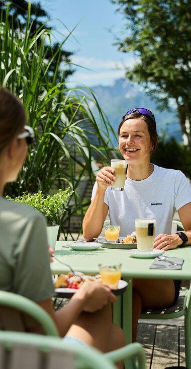 Two women enjoying a meal and drinks outdoors on a terrace during summer, surrounded by greenery and blue sky. | © Kleinwalsertal Tourismus eGen • Photographer: Oliver Farys