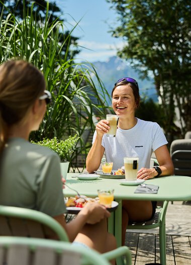 Zwei Frauen genießen eine Mahlzeit und Getränke im Sommer auf einer Terrasse, umgeben von grünen Pflanzen und blauer Himmel. | © Kleinwalsertal Tourismus eGen • Fotograf: Oliver Farys