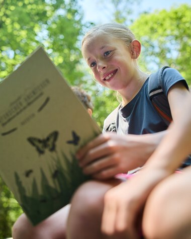 A smiling girl reads a nature brochure in a lush green forest during summer 2024. | © Kleinwalsertal Tourismus eGen • Photographer: Oliver Farys