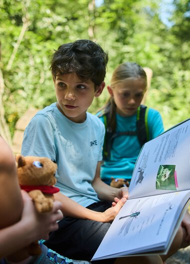 Three children outdoors, one reading a book, another holding a plush toy, during a nature exploration adventure in summer 2024 in Kleinwalsertal. | © Kleinwalsertal Tourismus eGen • Photographer: Oliver Farys