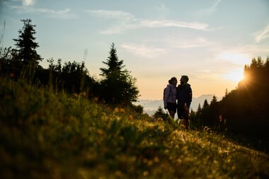 Two people stand during sunrise in a landscape with trees and hills, enjoying nature in Kleinwalsertal. | © Kleinwalsertal Tourismus eGen • Photographer: Oliver Farys