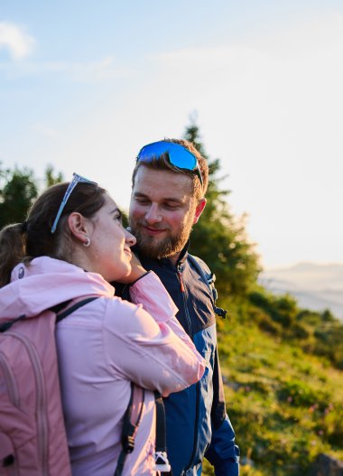A couple hiking at sunrise in Kleinwalsertal, surrounded by green mountains and trees, smiling and facing each other. | © Kleinwalsertal Tourismus eGen • Photographer: Oliver Farys