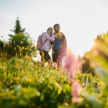A couple hiking in a blooming field at sunrise in Kleinwalsertal, surrounded by nature and trees. | © Kleinwalsertal Tourismus eGen • Photographer: Oliver Farys