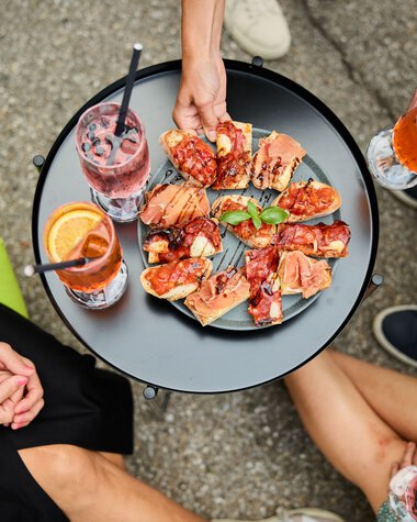 Three people sit around a small black table in summer, drinking beverages with lemon slices and sharing a platter of various warm pizza slices. | © Kleinwalsertal Tourismus eGen • Photographer: Oliver Farys