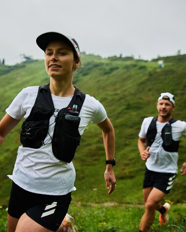 Two trail runners, a woman and a man, running on a green hillside in Kleinwalsertal during summer. Both wear running gear and vests. | © Kleinwalsertal Tourismus eGen • Photographer: Oliver Farys
