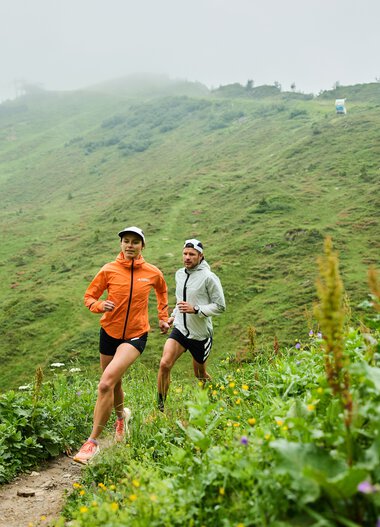 Two runners in athletic clothing running on a green trail in the mountainous Kleinwalsertal region, surrounded by plants and flowers. | © Kleinwalsertal Tourismus eGen • Photographer: Oliver Farys