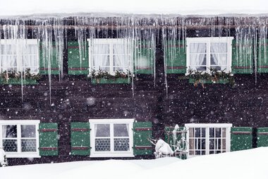 Snow-covered house with green shutters and icicles in winter during heavy snowfall. | © Kleinwalsertal Tourismus eGen • Photographer: Oliver Farys