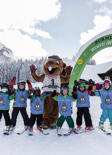 Children in ski gear and a mascot pose in the snow at a kindergarten ski course, under an archway reading 'Burmi Winter World' in a snowy mountain area. | © Kleinwalsertal Tourismus eGen • Photographer: Oliver Farys