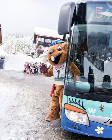 Mascot in winterwear next to a bus in snowy landscape, with children in the background, winter clothing. | © Kleinwalsertal Tourismus eGen • Photographer: Oliver Farys