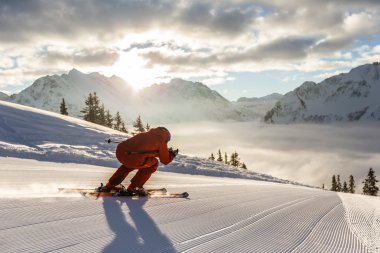Skifahrer in roter Kleidung fährt auf frisch präparierter Piste in den Bergen, Sonnenlicht und Wolken am Himmel. | ©  Kleinwalsertal Tourismus eGen • Fotograf: Oliver Farys