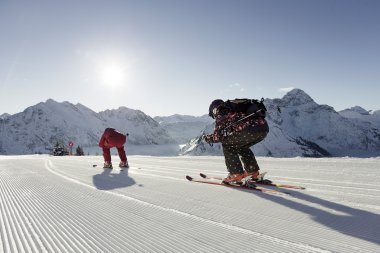 Two skiers on freshly groomed snow in the mountains, with the sun and snow-covered peaks in the background. | © Kleinwalsertal Tourismus eGen • Photographer: Oliver Farys