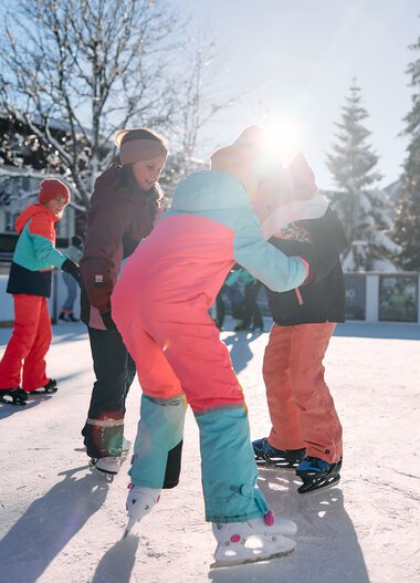 Children ice skating on an outdoor rink in winter, surrounded by snow and trees with a building in the background. | © Kleinwalsertal Tourismus eGen • Photographer: Oliver Farys